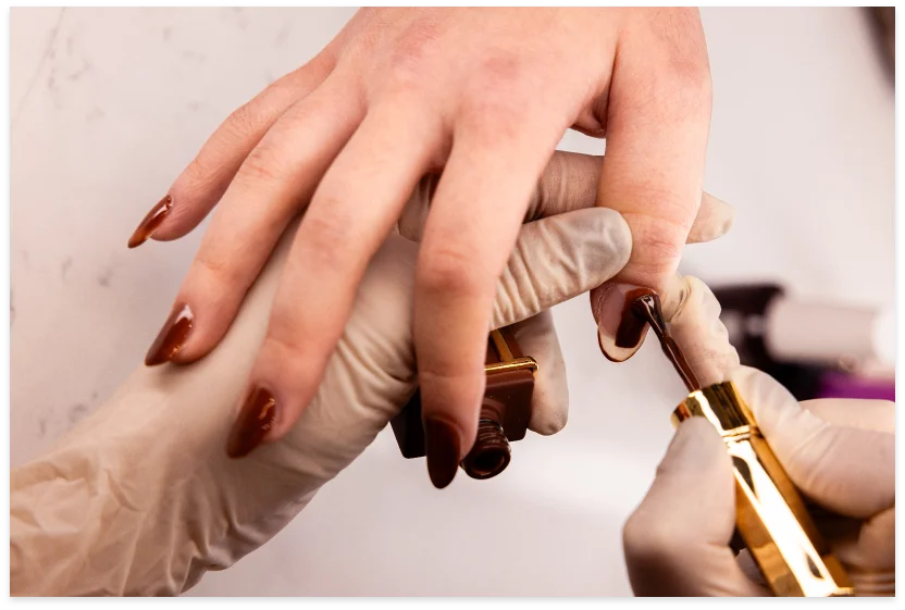 Chocolate-brown polish being applied to long almond nails beside a gold-capped bottle